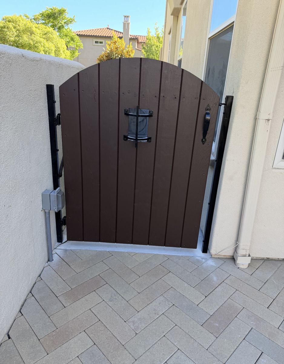 Arched dark-brown wood side gate with black metal posts, speakeasy grille and handle over herringbone pavers